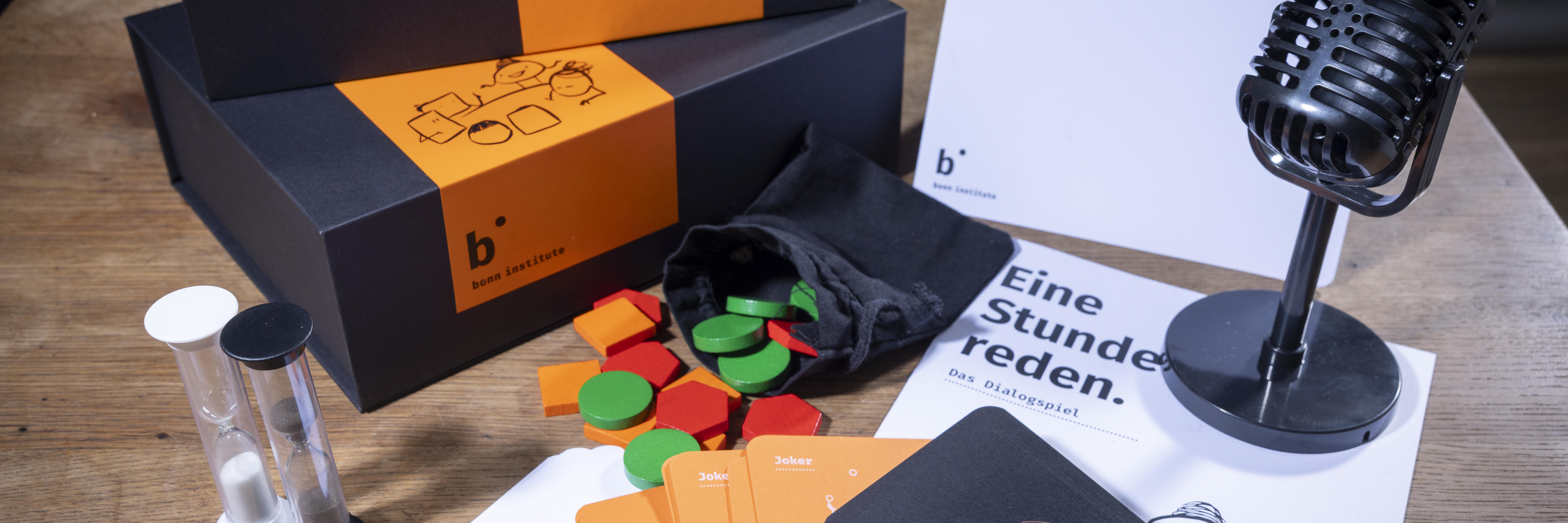 Das Spielset von "Eine Stunde reden" liegt ausgebreitet auf einem Tisch. Zu sehen sind der Spielkarton, eine Sanduhr, ein Mikrofon, eine Spielanleitung und verschiedene Spielteile- und karten.
